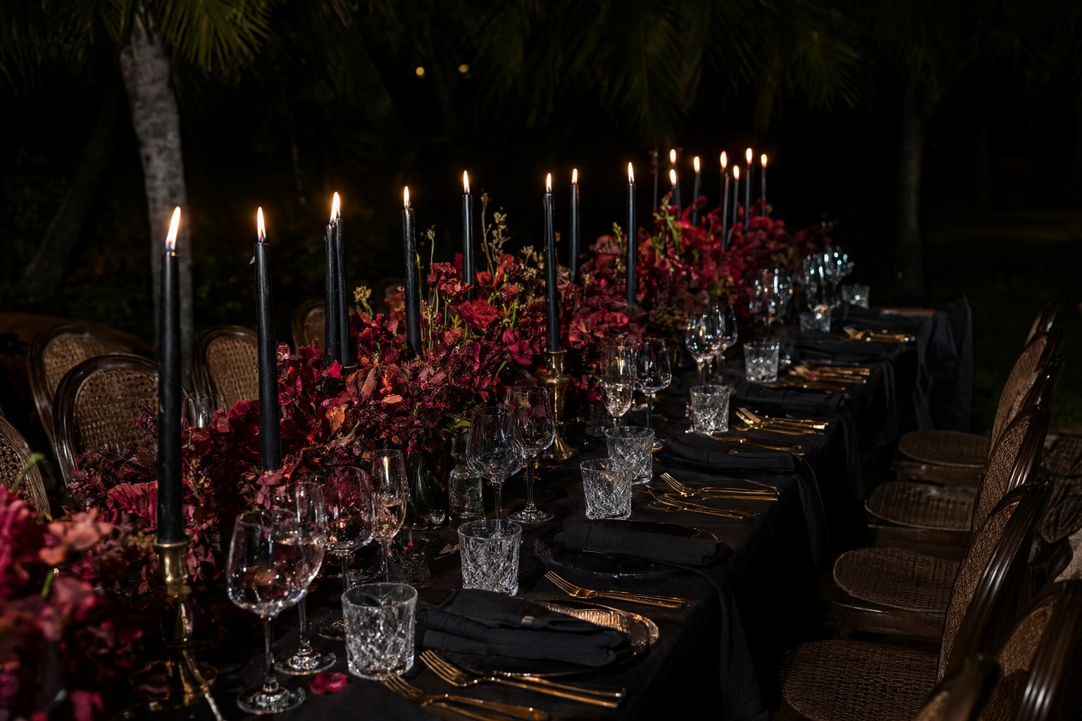 A long candlelit dinner table with dark florals under palms