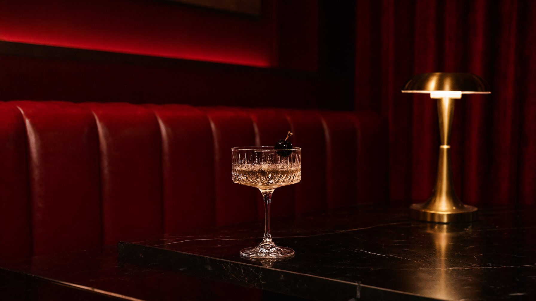 A private lounge in deep red light, a single cherry-topped coupe on a black marble bar